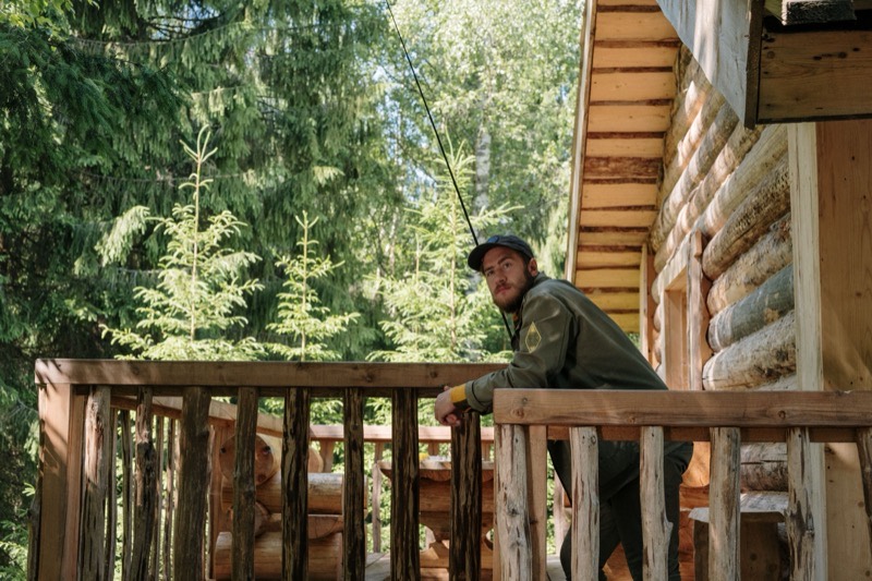 A man alone on the deck of a timber cabin, leaning on the rail and looking out at the trees.